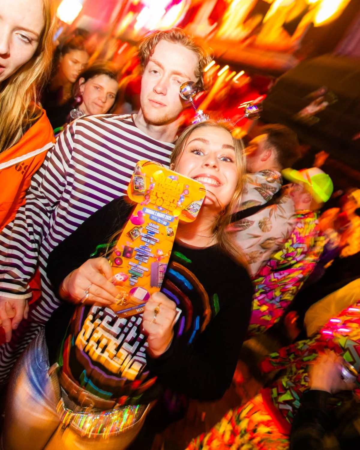 Lachende vrouw houdt een mega das naast haar gezicht, tussen vrienden in een volle kroeg