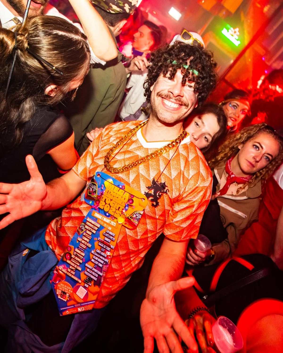 Man met krullenpruik en oranje voetbalshirt toont zijn mega das in een volle kroeg